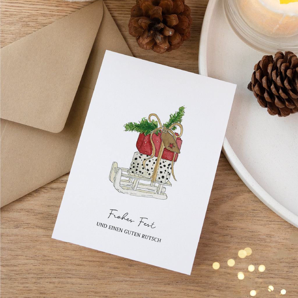 Festliche Weihnachtskarte mit Schlitten und Geschenken als handgemaltes Aquarell Motiv neben Umschlag und Zapfen.