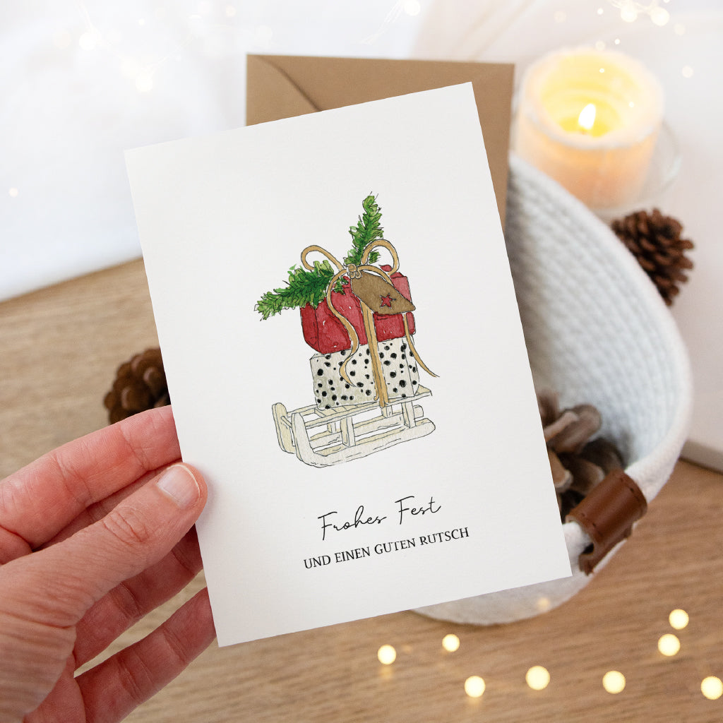 Hand hält eine weihnachtliche Klappkarte mit Schlitten Aquarell Motiv über einem weißen Korb mit Tannenzapfen.
