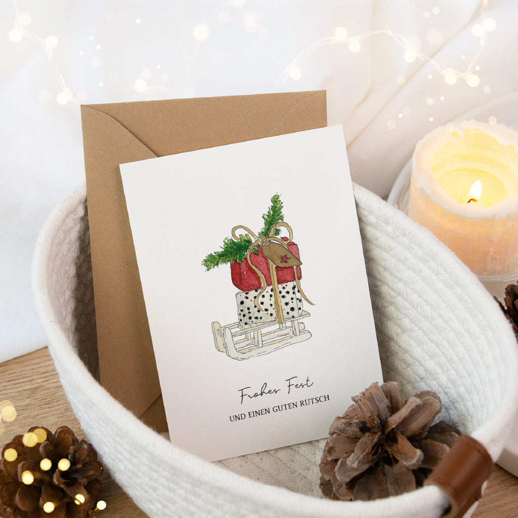 Weihnachtspost Karte mit Schlitten Aquarell Motiv und Umschlag in einem weißen Stoffkorb mit Kerze und Zapfen.