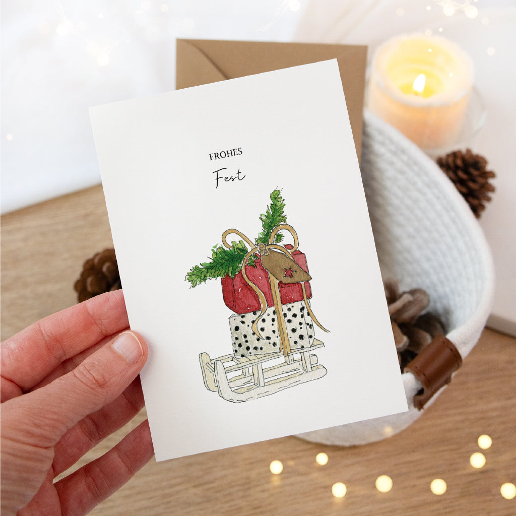 Hand hält die Weihnachtskarte mit buntem Schlitten Aquarell Motiv über einen weißen Korb im warmen Kerzenlicht.