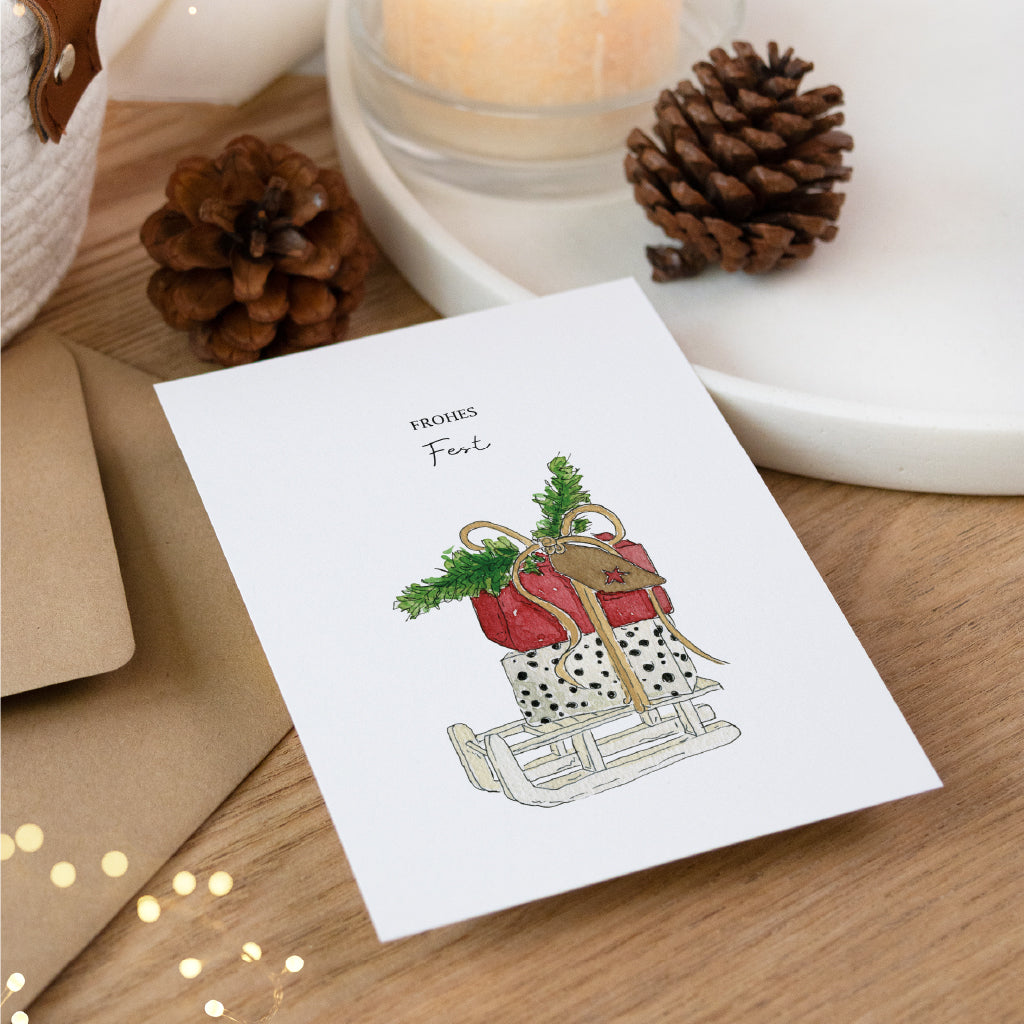 Hochwertige Weihnachtspost mit Schlitten Aquarell Motiv auf Holztisch dekoriert mit Lichterkette und Umschlag.