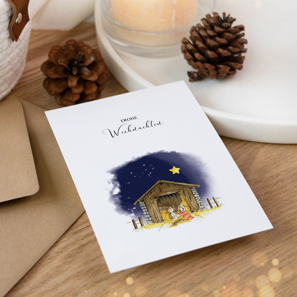 Aquarell-Weihnachtskarte mit liebevoller Darstellung der Heiligen Familie vor dem Stall, eingerahmt von winterlicher Deko.