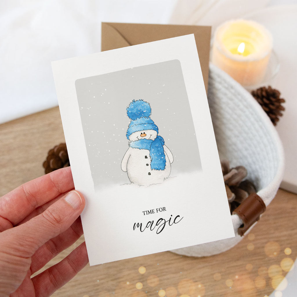 Weihnachtskarte als Klappkarte mit Aquarell-Schneemann in Blau, winterlich illustriert, wird von einer Hand gehalten – perfekte Karte für die Weihnachtszeit.
