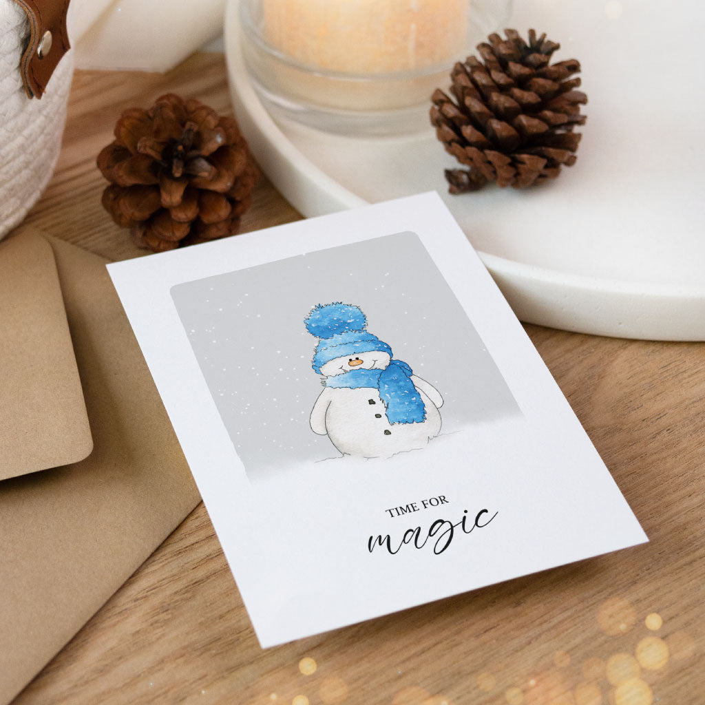 Liebevoll gestaltete Winterkarte mit Schneemann-Aquarell und Aufschrift „Time for magic“, auf hellem Holztisch mit Kerze und Umschlag arrangiert.