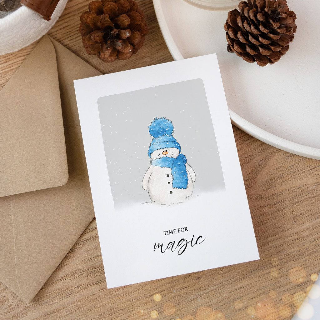 Klappkarte mit niedlichem Schneemann in Aquarelltechnik, stimmungsvoll inszeniert mit braunem Briefumschlag und winterlicher Dekoration.