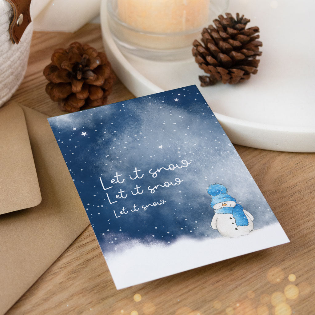Winterliche Postkarte mit Schneemann und Schneeflocken vor dunklem Aquarellhimmel, zusammen mit Umschlag und Dekoration für Geschenkverpackungen abgebildet.