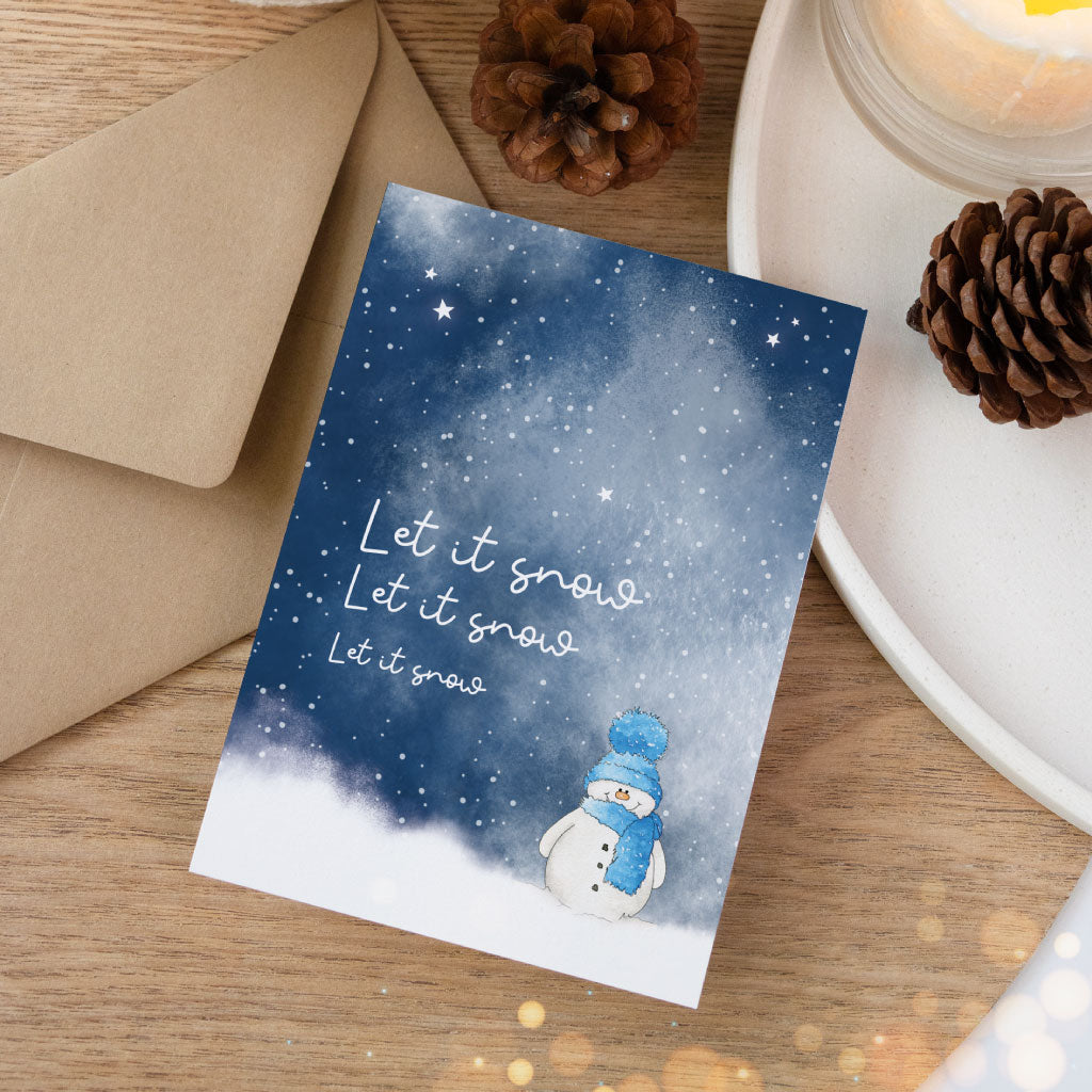 Handgemalte Weihnachtskarte im Aquarell-Stil mit kleinem Schneemann und Schneeflocken, passend zu natürlichen Geschenkverpackungen mit Tannenzapfen und Kerzenlicht.