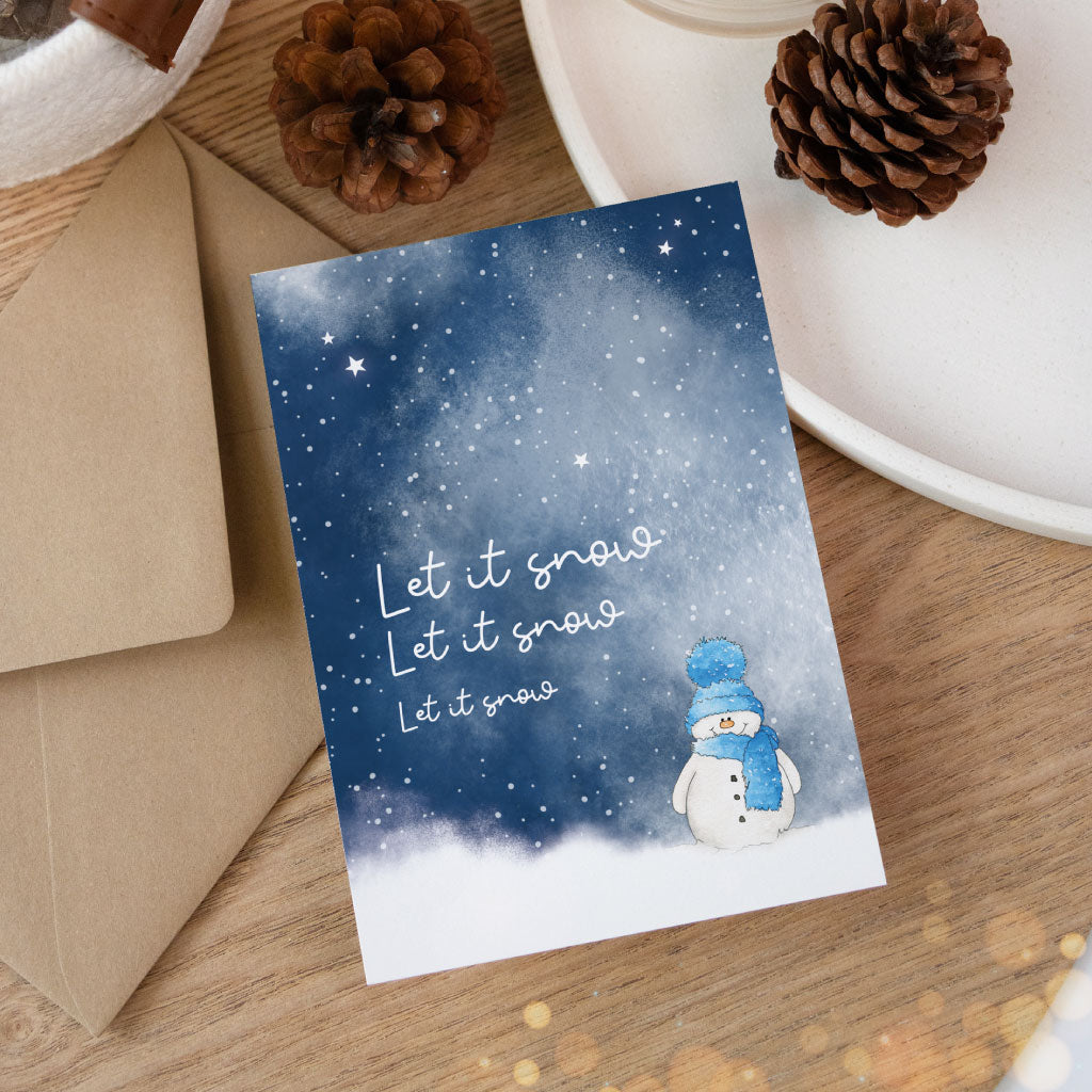 Stimmungsvolles Kartenmotiv mit Aquarell-Schneemann und „Let it snow“-Schriftzug, arrangiert auf Holzuntergrund mit Kraftpapierumschlag und weihnachtlicher Deko.