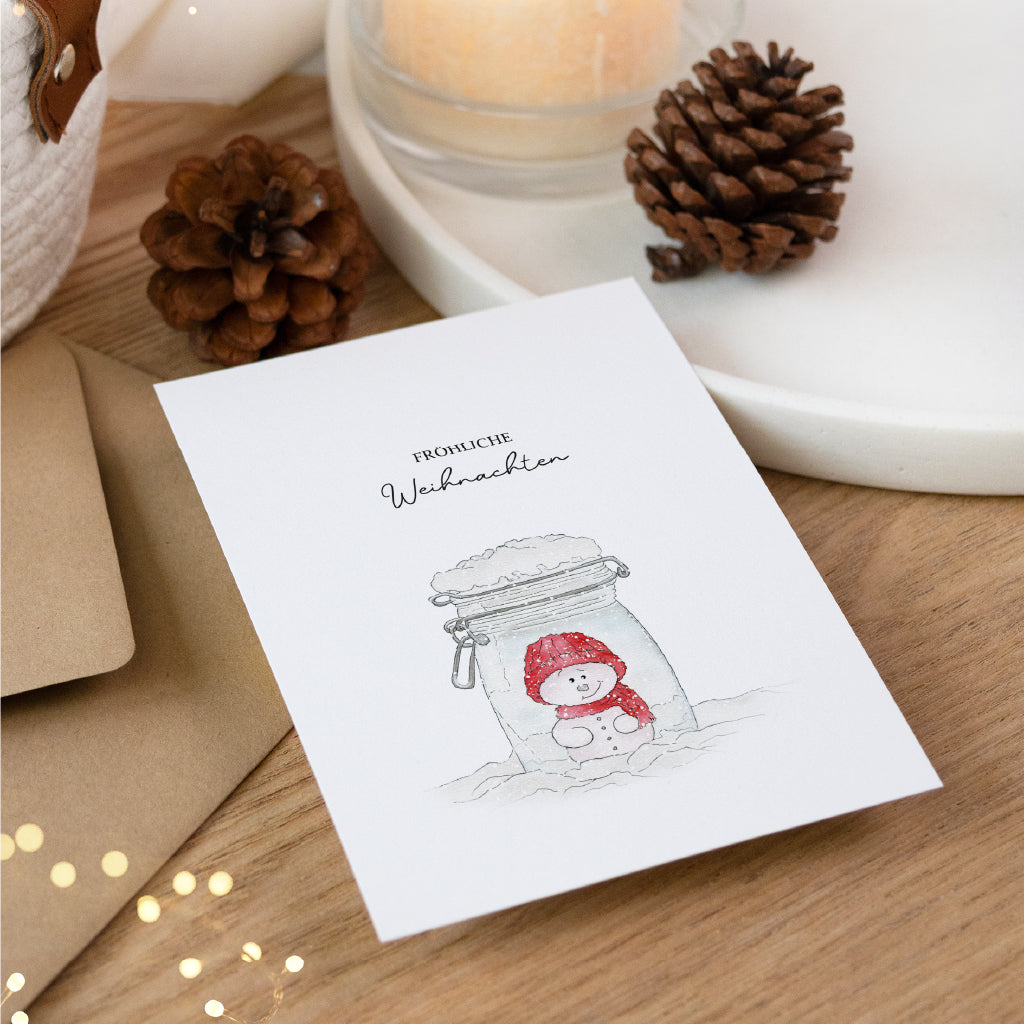 Detailansicht der Postkarte mit handgemaltem Schneemann Aquarell Motiv neben funkelnden Lichtern und Tannenzapfen.