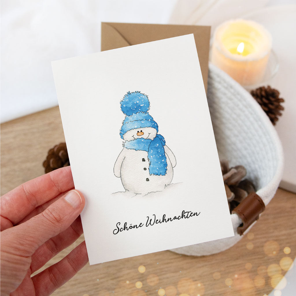 Postkarte mit Schneemann-Aquarellmotiv und Schriftzug „Schöne Weihnachten“, gehalten in der Hand vor winterlicher Dekoration – perfekt als Weihnachtskarte oder Geschenkbeileger.