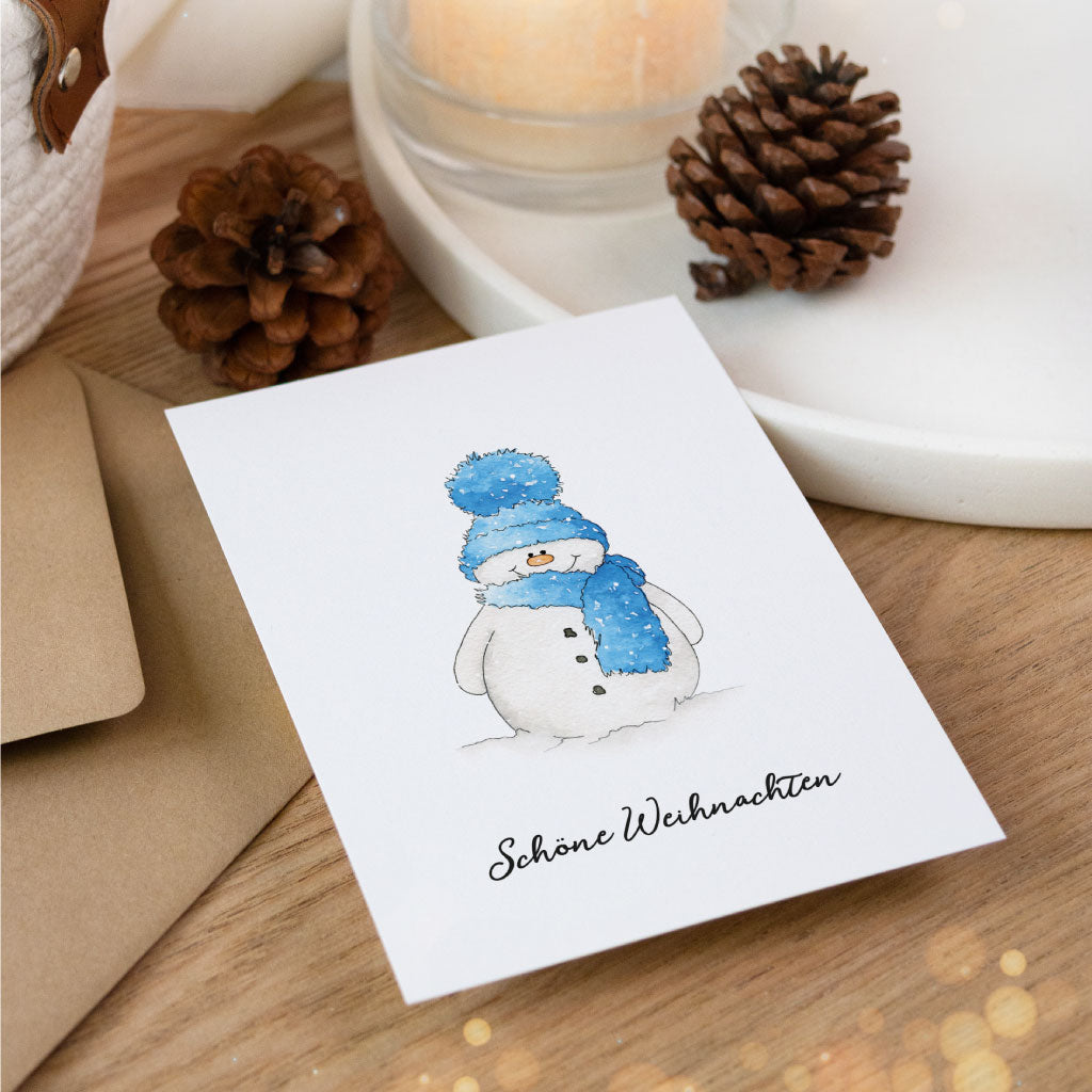 Weihnachtskarte mit Aquarell-Schneemann in blauer Mütze und Schal, auf Holztisch mit Kerze, Umschlag und Tannenzapfen – stilvolle Postkarte zum Weihnachtsgeschenke verpacken.
