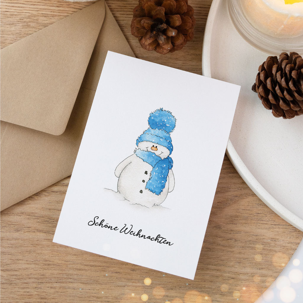 Handgemalte Aquarellkarte mit Schneemann-Motiv und Aufschrift „Schöne Weihnachten“, dekorativ arrangiert neben Kerze, Umschlag und Tannenzapfen.