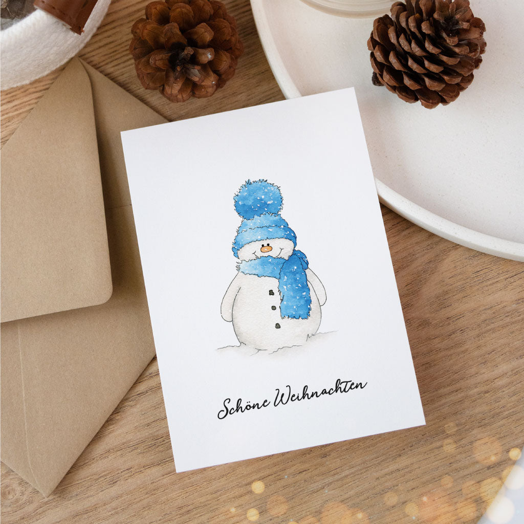 Aquarell-Weihnachtskarte mit Schneemannmotiv auf Holztisch, kombiniert mit braunem Umschlag und weihnachtlicher Deko – ideal als Postkarte zu Weihnachten.
