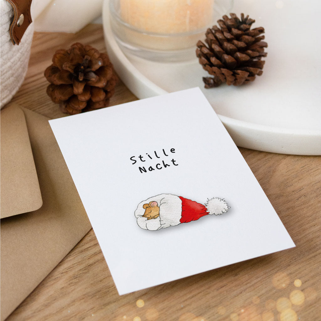 Weihnachtlich inszenierte Aquarell-Postkarte mit Motiv einer ruhenden Maus in einer roten Mütze und dem Spruch „Stille Nacht“, arrangiert mit Umschlag und Deko.