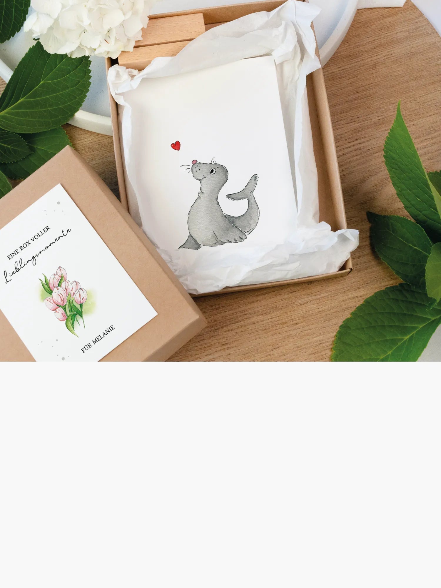 Geöffnete Geschenkbox mit Aquarellkarte "Eine Box voller Lieblingsmomente" und Grußkarte mit Robben-Motiv