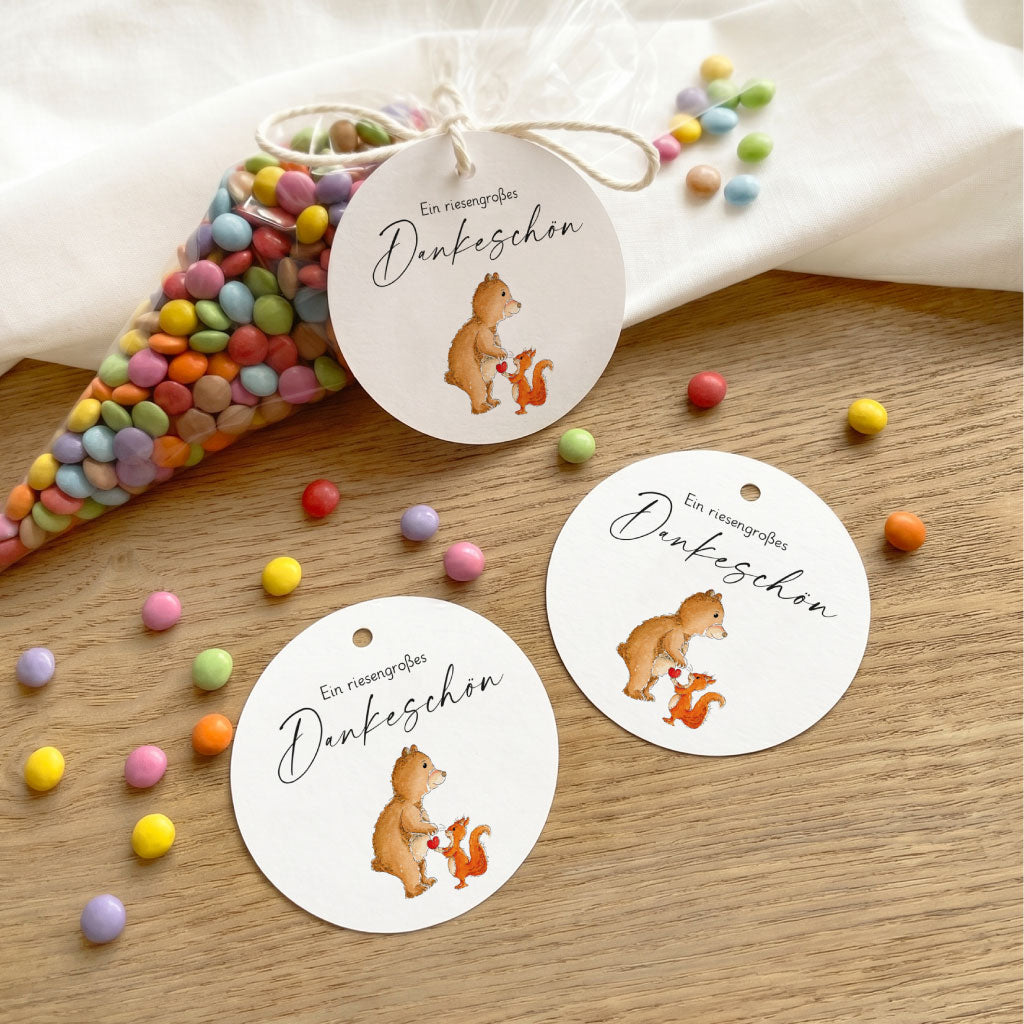 Drei Geschenkanhänger mit Bär und Eichhörnchen Aquarell Motiv neben einer Spitztüte mit bunten Schokolinsen.