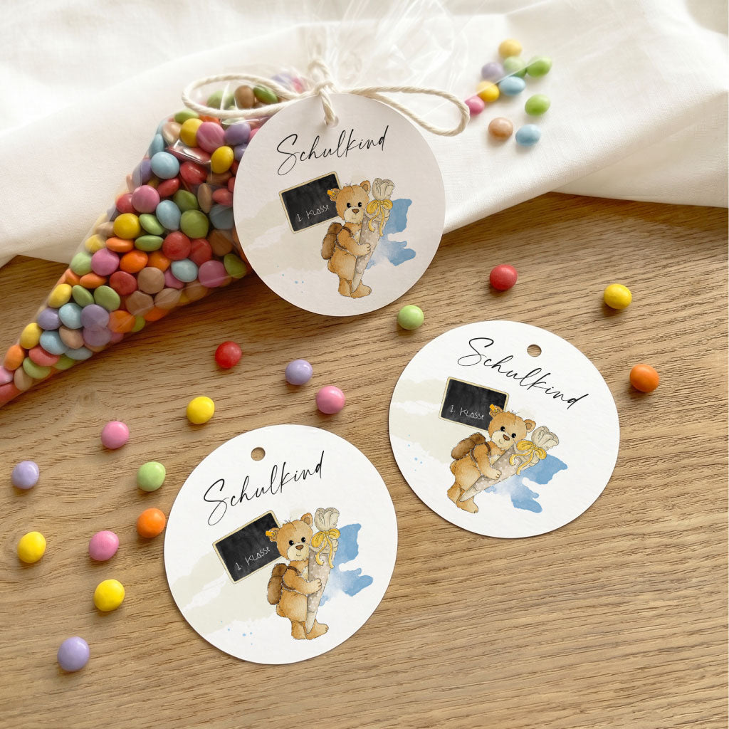 Drei runde Geschenkanhänger zur Einschulung mit Teddy Aquarell Motiv neben einer Spitztüte mit bunten Schokolinsen.