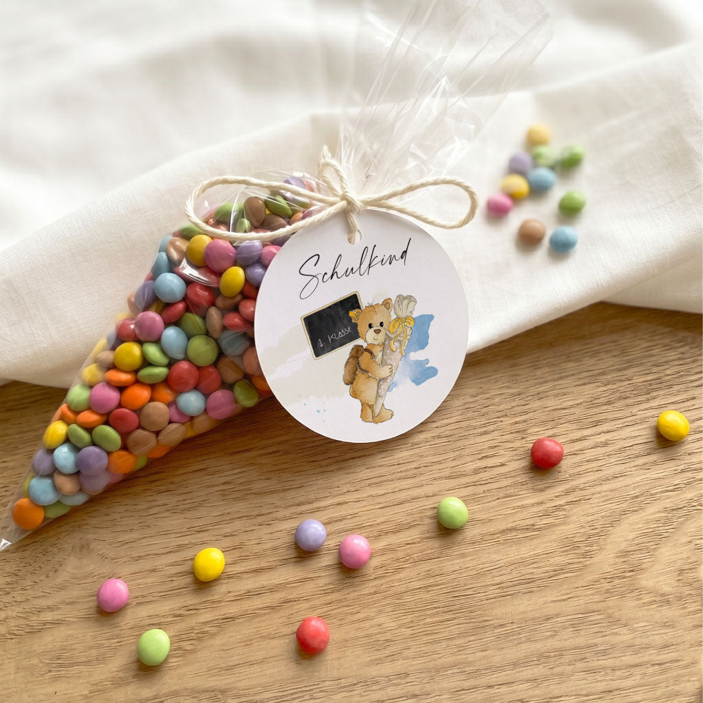 DIY Deko für die Einschulungsparty: Anhänger mit Teddy Aquarell Motiv an einer transparenten Tüte mit Süßigkeiten.