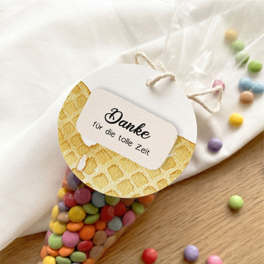 Detailansicht des runden Anhängers mit Eiswaffel Motiv an einer Tüte mit bunten Schokolinsen auf hellem Stoff.