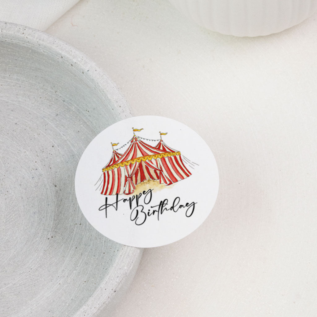 Runder Sticker mit Zirkuszelt zum Geburtstag und Schriftzug „Happy Birthday“ auf heller Oberfläche, als Geburtstagsgeschenk-Verpackung und Dekoration