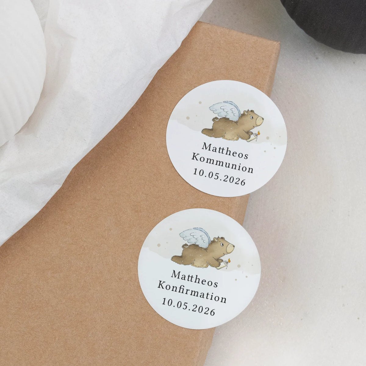 Zwei runde Aufkleber zur Kommunion und Konfirmation mit personalisiertem Namen und Datum auf Kraftpapierpaket, Aquarell Motiv fliegender Engelbär