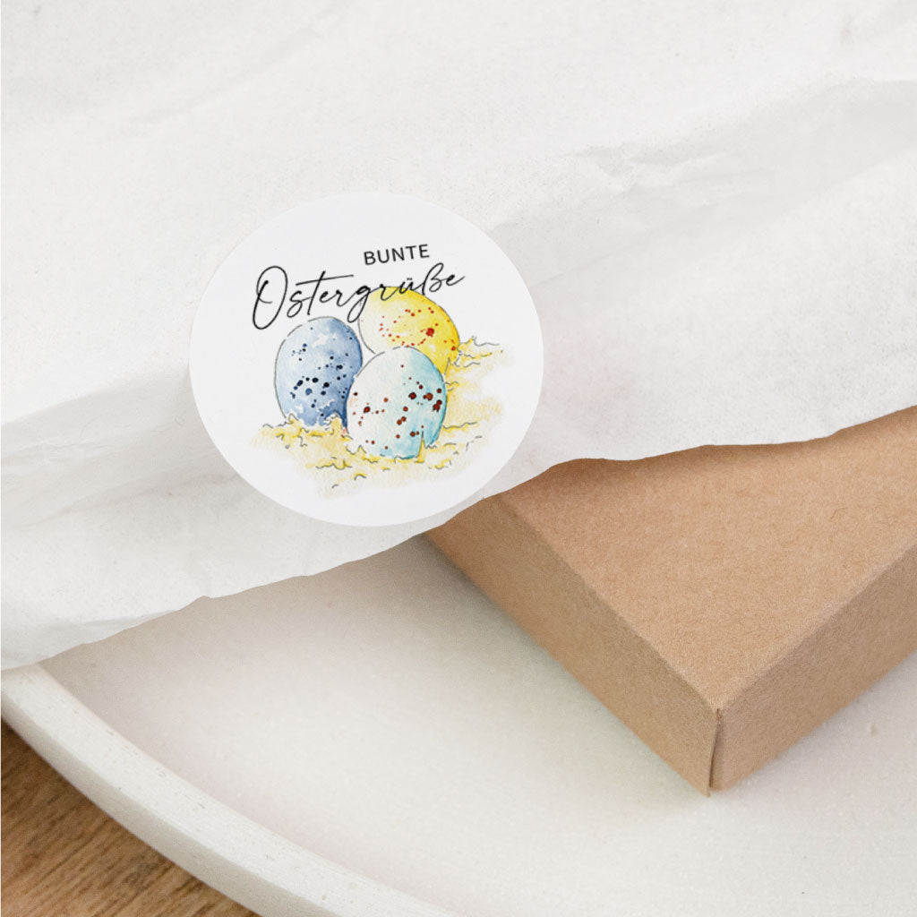 Rundes Sticker-Motiv „Bunte Ostergrüße“ mit drei Ostereiern auf Seidenpapier und Kraftkarton, osteraufkleber für geschenkverpackung zu ostern.
