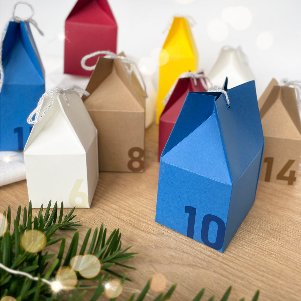 Farbenfrohes DIY Bastelset mit kleinen Adventskalender-Häusern aus Karton in Blau, Weiß, Natur, Rot und Gelb – ideal für kreative Adventsdekoration.