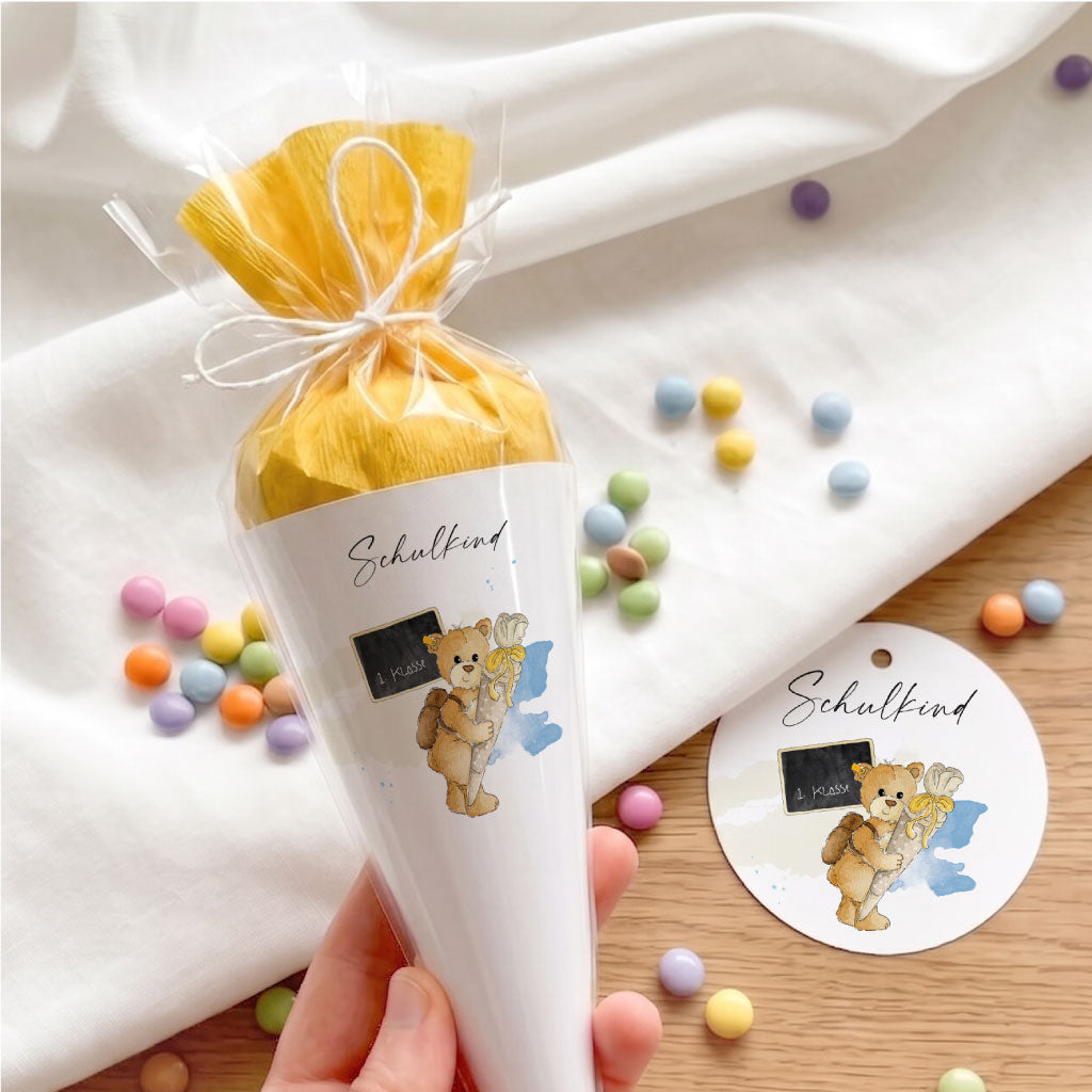 Kleine DIY Schultüte in der Hand und passender runder Geschenkanhänger mit Teddy Aquarell Motiv zur Einschulung.
