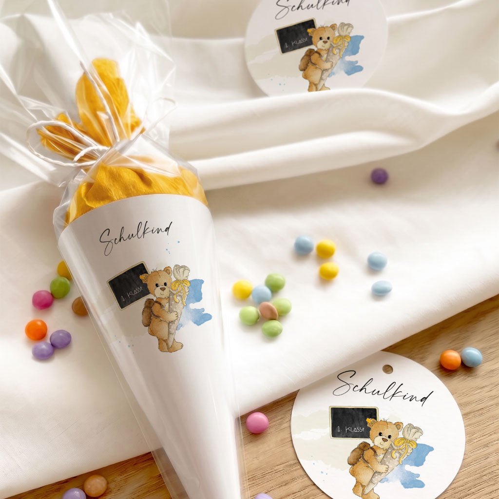 Mini Schultüte verpackt in Folie neben runden Anhängern mit Teddy Aquarell Motiv als Deko zur Einschulung.