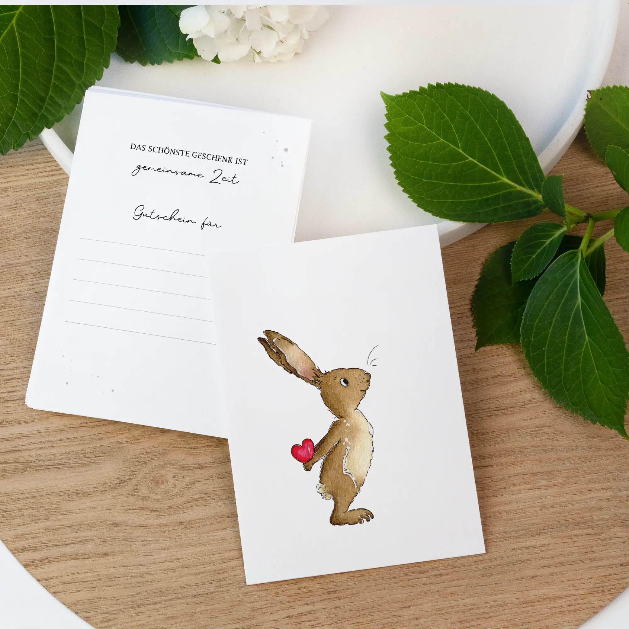 Zwei weiße Gutscheinkarten auf einem Holztisch. Vorne ein handgemaltes Hase Aquarell Motiv und hinten Linien zum Ausfüllen.