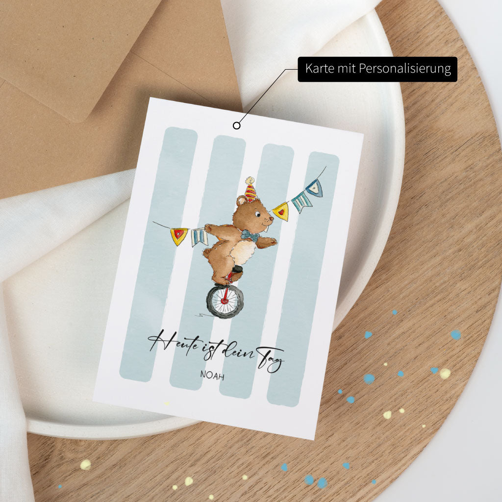 Geburtstagskarte mit Aquarell-Illustration eines Zirkusbären auf Einrad, optional mit Personalisierung, auf Holzplatte mit dezentem Konfetti arrangiert.