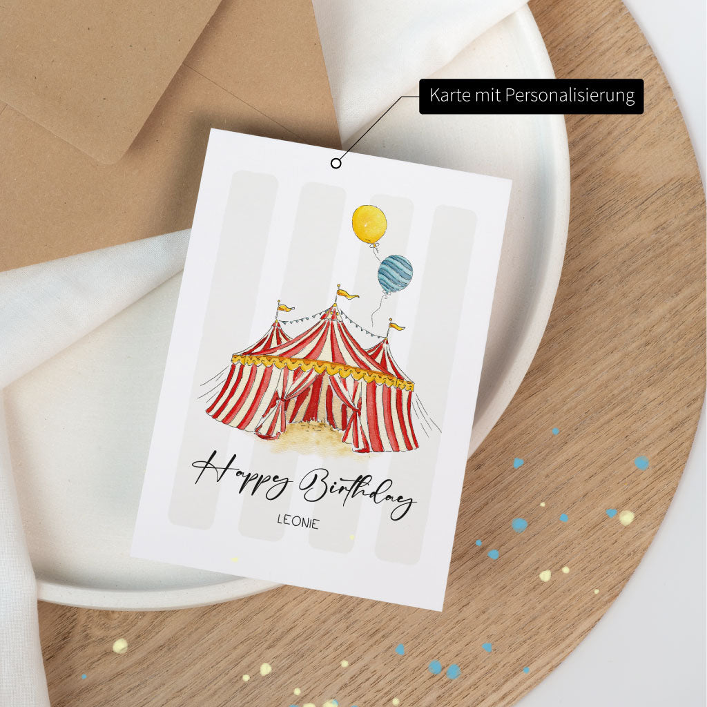 Geburtstagskarte mit Aquarell-Zirkuszelt und personalisierbarem Namensfeld, auf Holzplatte mit farbigen Sprenkeln dekorativ fotografiert.