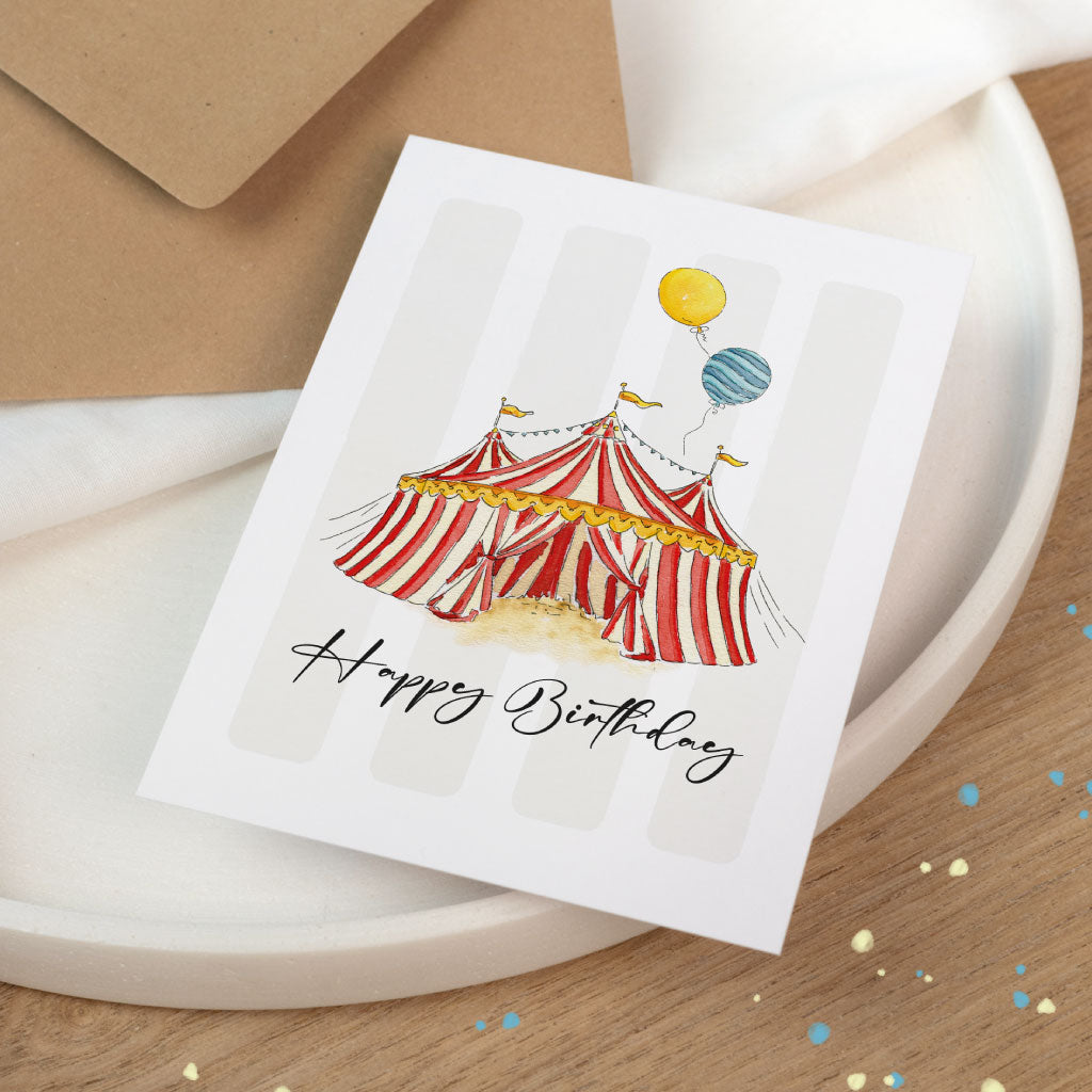 Stimmungsvolle Szene mit Geburtstagskarte im Zirkusdesign ohne Personalisierung, liebevoll illustriert in Aquarell, daneben ein brauner Umschlag.
