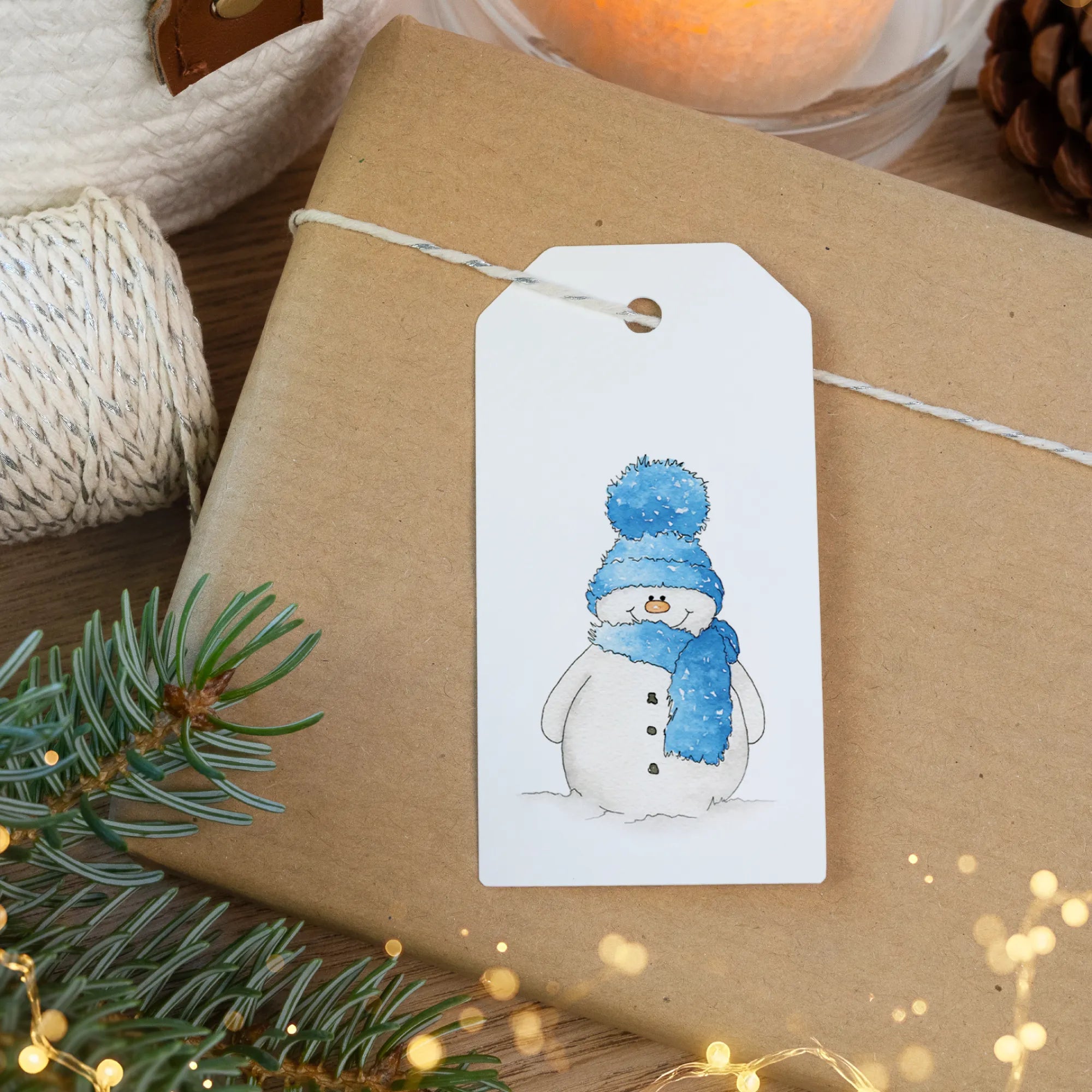 Geschenkanhänger mit fröhlichem Schneemann Aquarell Motiv mit blauer Mütze auf rustikaler Geschenkverpackung.