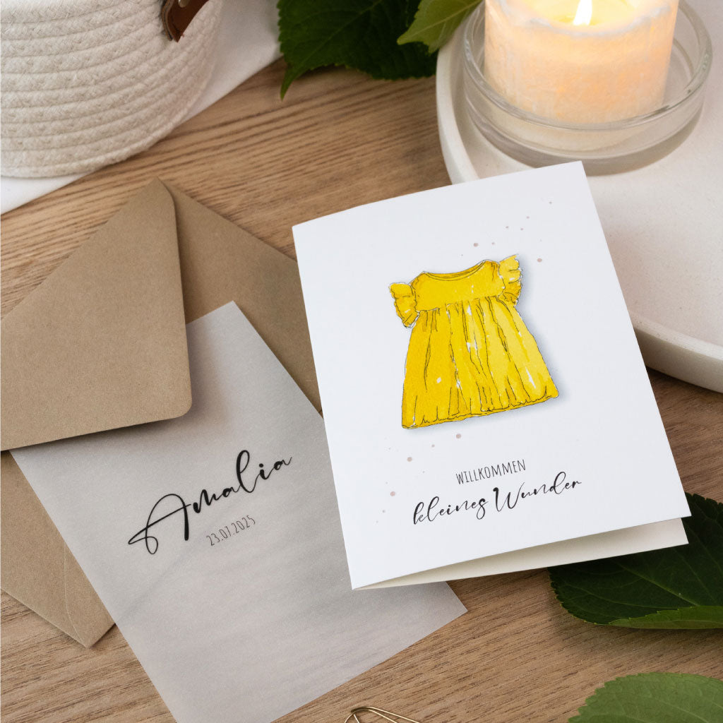 Personalisierte Glückwunschkarte zur Geburt für Mädchen mit handgemaltem Aquarellmotiv eines gelben Kleids und Transparentdeckblatt mit Name und Geburtsdatum.