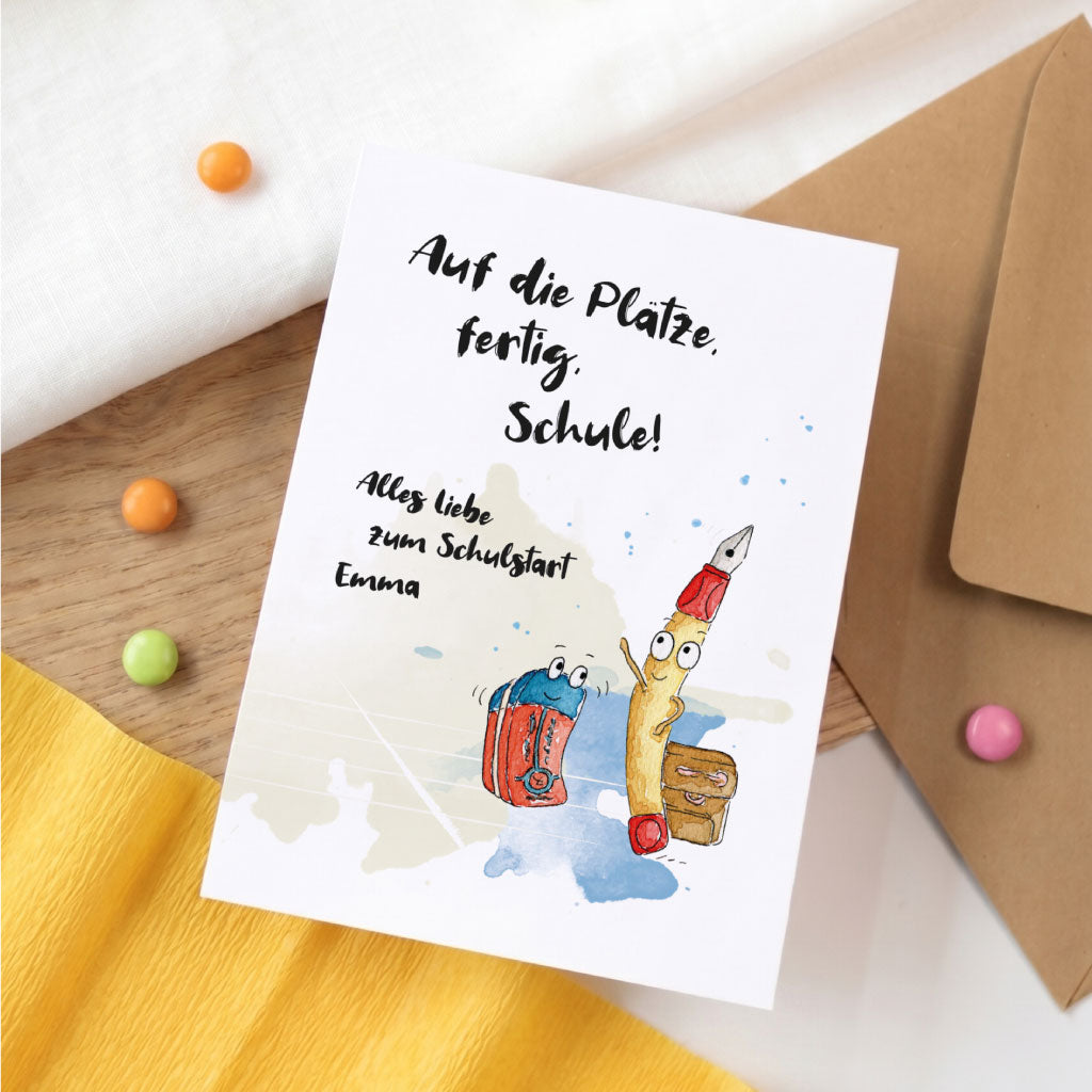 Karte zur Einschulung mit Stifte Aquarell Motiv und Name Emma auf Holztisch neben bunten Schokolinsen.