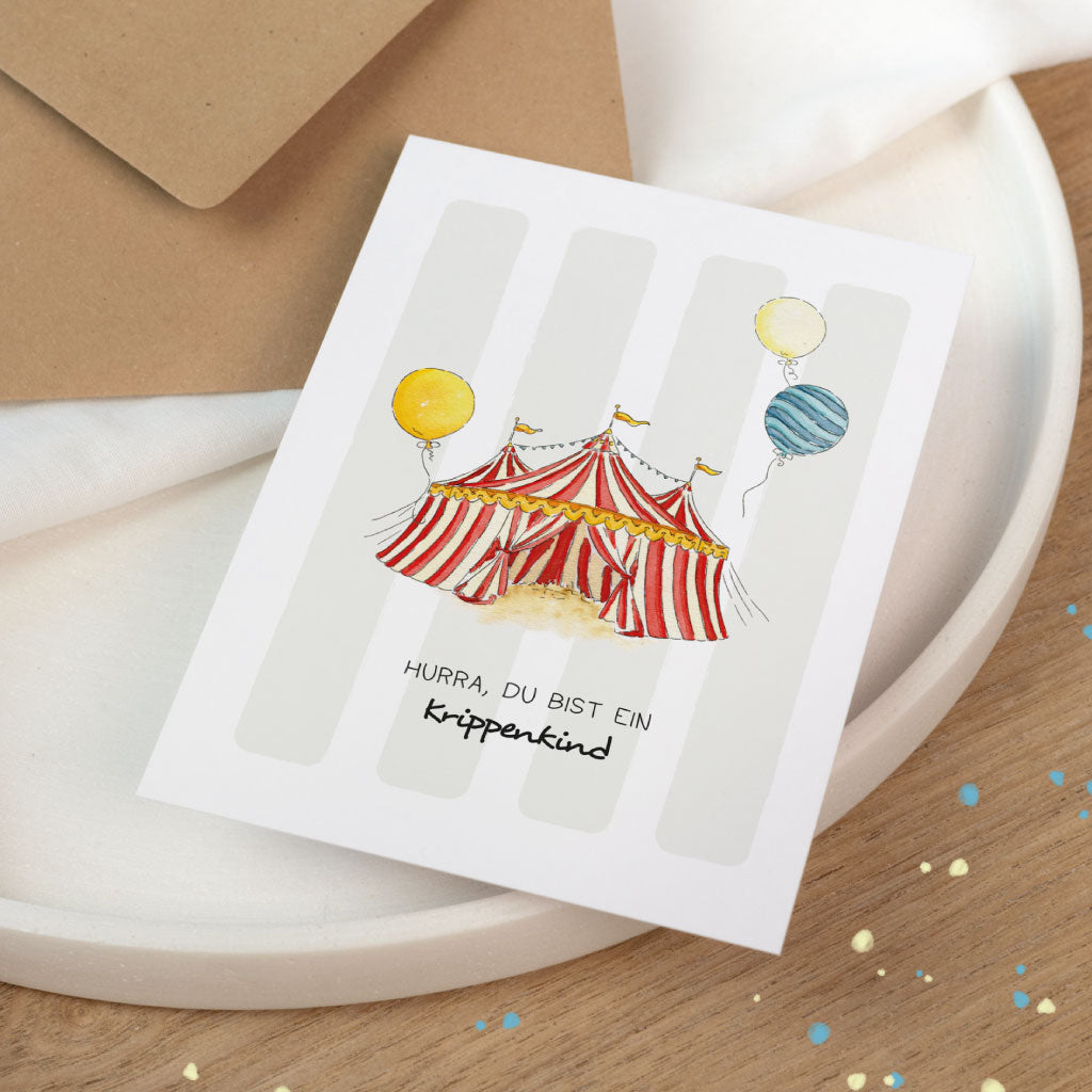 Schräg fotografierte Karte zum Krippe-Beginn: Zirkus-Aquarellmotiv mit Ballons und Schriftzug „Hurra, du bist ein Krippenkind“