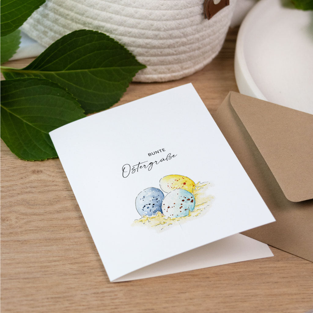 Handgemalte Osterkarte als Klappkarte mit bunten Ostereiern auf Stroh im Aquarell Stil und dem Text bunte Ostergrüße.