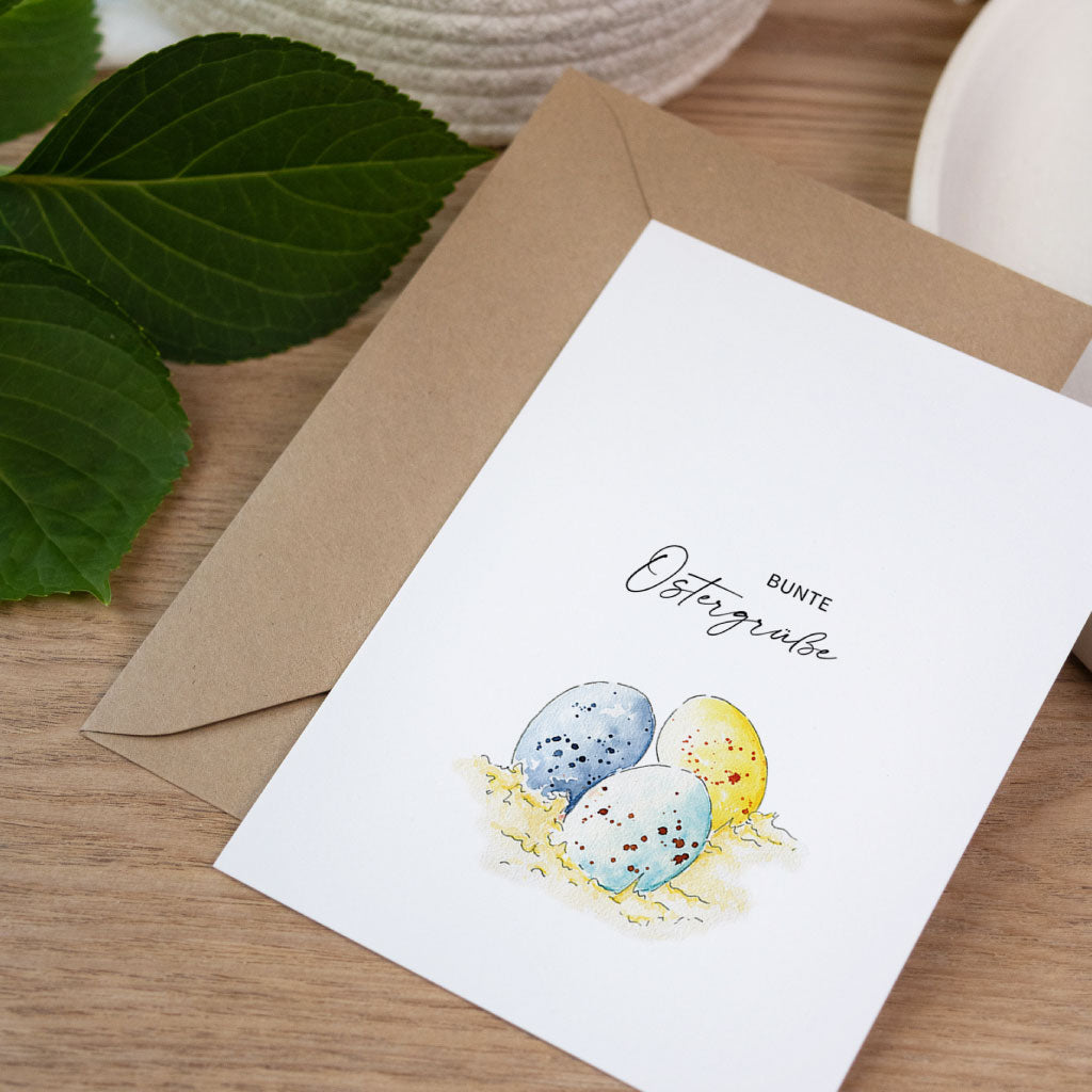 Osterkarte im Aquarell Stil mit bunten Ostereiern auf Stroh und Schriftzug bunte Ostergrüße.