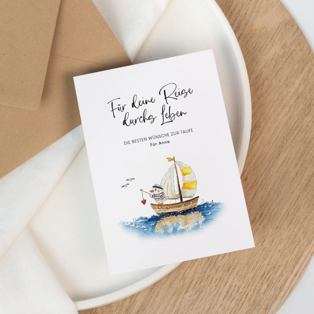 Taufkarte mit handgemaltem Schiff Aquarell Motiv und kleinem Matrosen auf dem Meer neben einem Kraftpapier Umschlag.