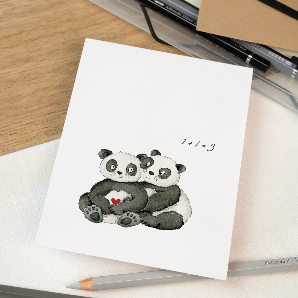 Karte zur Verkündung einer Schwangerschaft mit handgemalten Pandas und rotem Herz, Aufschrift 1+1=3, auf Stoffuntergrund