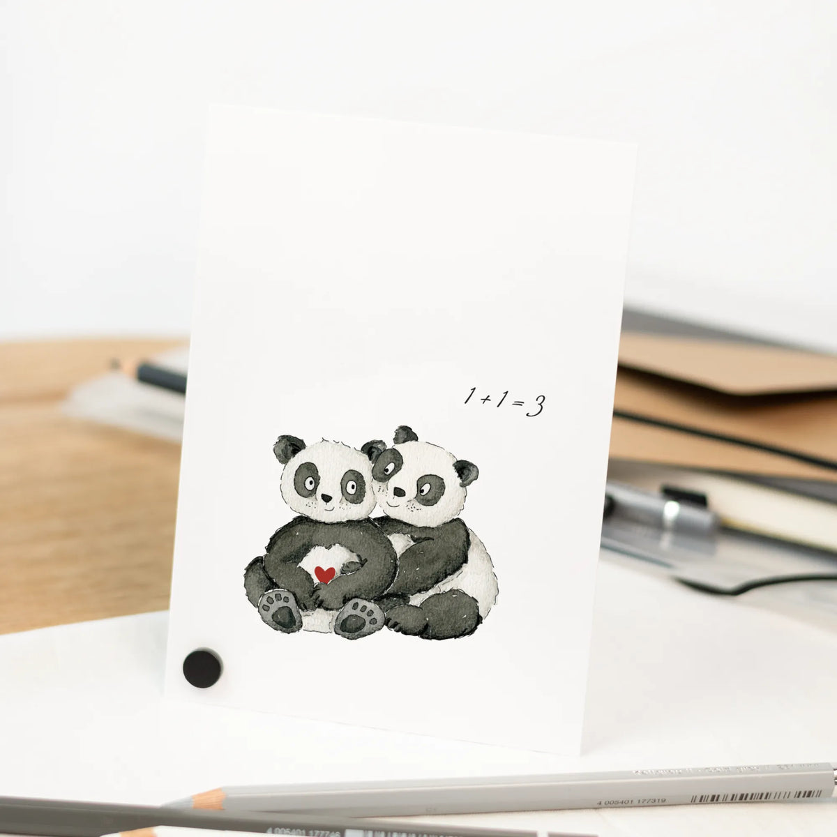 Schwangerschaftskarte mit handgemalten Aquarell Pandas und Aufschrift 1+1=3, stehend vor weißem Hintergrund mit Zeichenmaterial