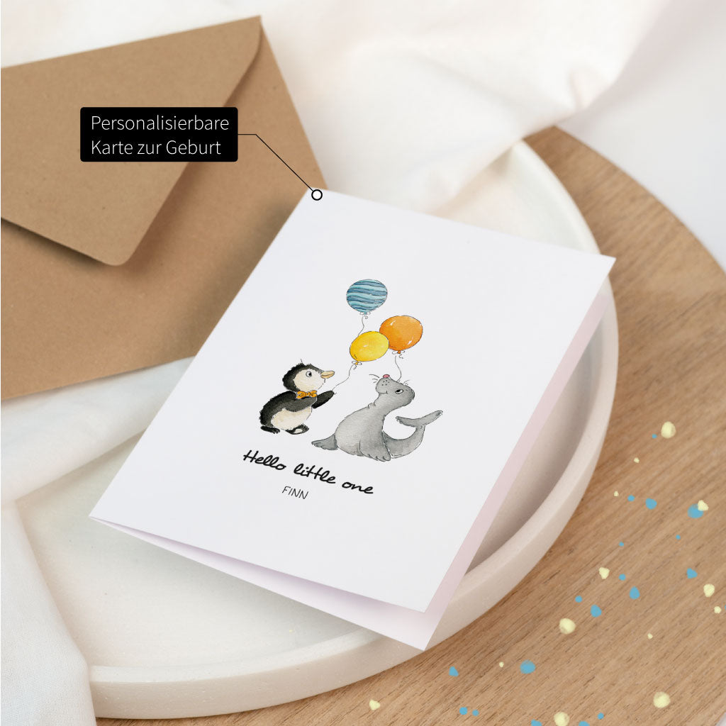 Gefaltete Glückwunschkarte zur Geburt eines Babys, mit Name personalisierbar: Pinguin und Seehund mit Luftballons auf weißer Karte, daneben brauner Umschlag.