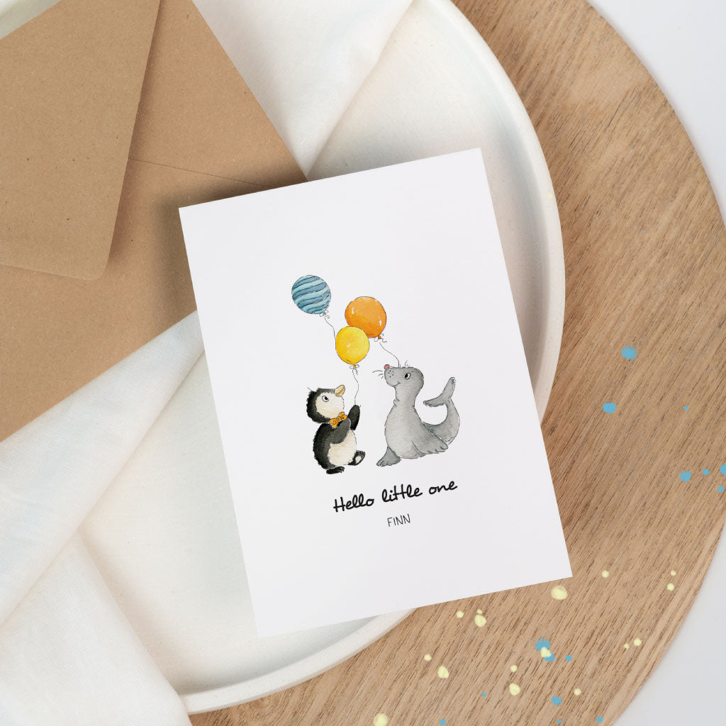 Produktfoto: Glückwuschkarte zur Geburt Baby auf Keramikteller und Holztisch, handgemaltes aquarell Motiv mit Pinguin, Seehund und drei Luftballons, Text „Hello little one“.