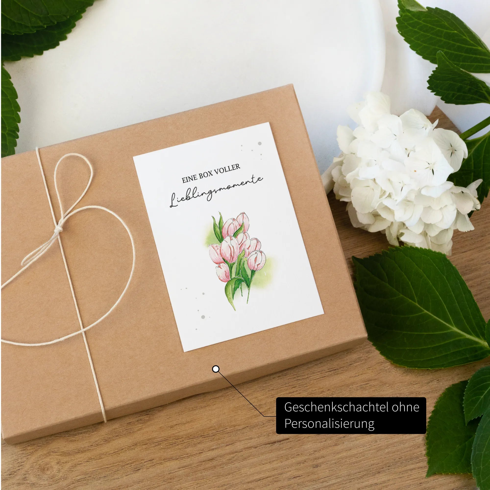 Geschenkbox gebunden mit weißem Band auf einem Holztisch. Das Deckblatt zeigt rosafarbene Tulpen ohne Personalisierung.