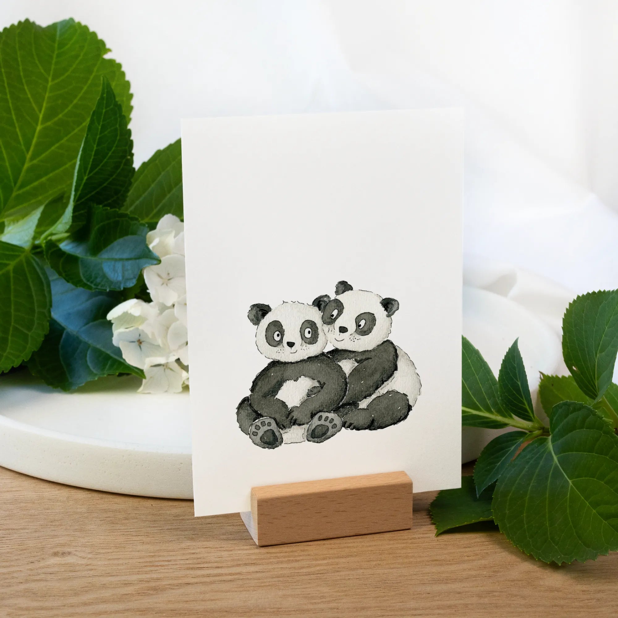 Eine weiße Gutscheinkarte steht in einem Holz Kartenhalter. Darauf ein Panda Aquarell Motiv in schwarz und weiß gemalt.
