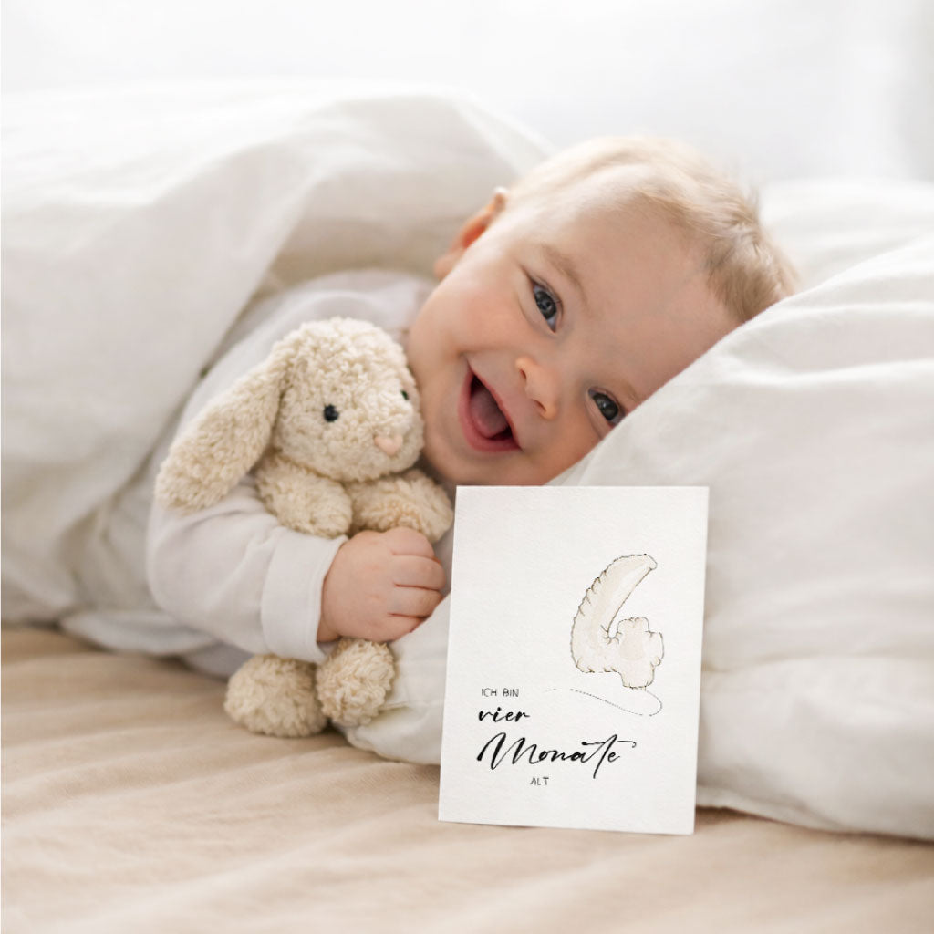 Lächelndes Baby im Bett mit Meilensteinkarte „vier Monate alt“ und Kuscheltier – liebevolle Erinnerungskarte aus dem Set fürs erste Babyjahr.