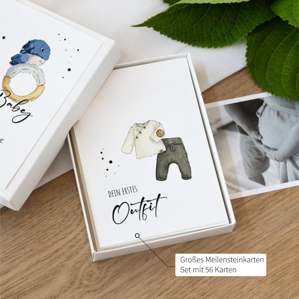 Geöffnete Box mit Aquarellkarten zur Schwangerschaft, obenauf liegt die Karte „Dein erstes Outfit“ mit Babykleidung in Naturtönen als handgemaltes Aquarell.