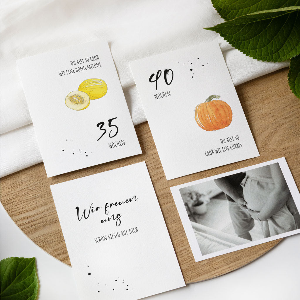 Drei Schwangerschaftskarten auf Holztisch: Honigmelone (35 Wochen), Kürbis (40 Wochen) und eine Karte mit Text „Wir freuen uns schon riesig auf dich“, alle mit liebevollen Aquarellmotiven.