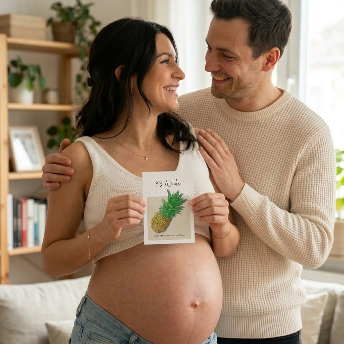 Frau hält Karte SSW 33 mit Ananas Motiv vor ihren Babybauch, ihr Partner steht hinter ihr und sie lächeln sich liebevoll an.